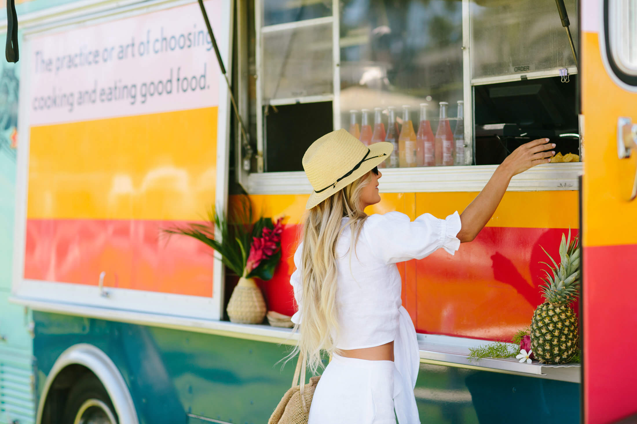 woman wearing a sunhat reaches her arm up to the window of the Aloha Trick Pony food truck