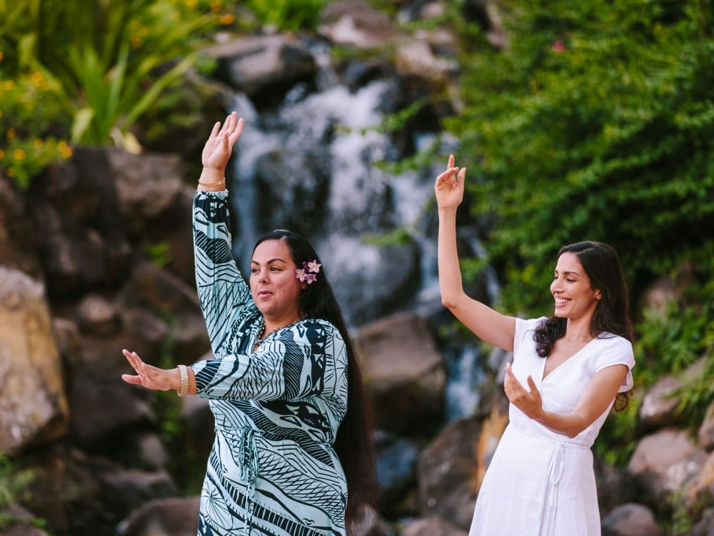 two women hold one hand above their head and one in front of them as they practice dancing in front of.a waterfall feature surrounded by foliage