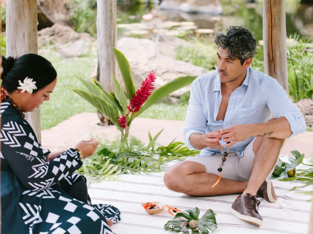 a woman in a blue striped dress with a white flower in her hair sits facing a man while weaving a bracelet