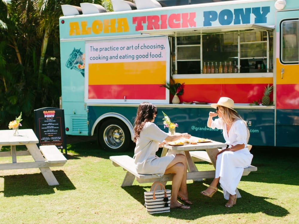 two people sit at a picnic table in front of the Aloha Trick Pony food truck