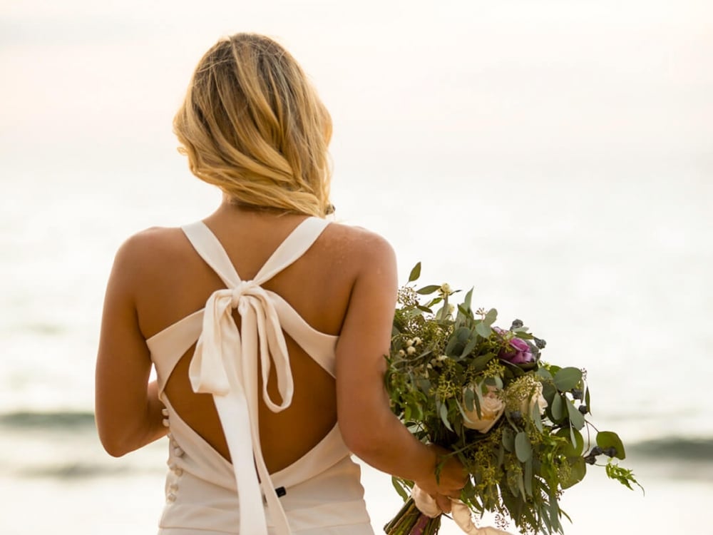 a bride in a white dress stands with a bouquet in her hand faces the ocean