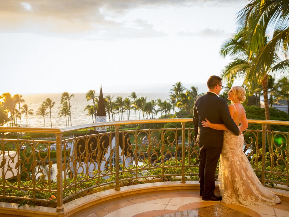a bride and groom stand on a balcony facing the sunset over Grand Wailea's chapel