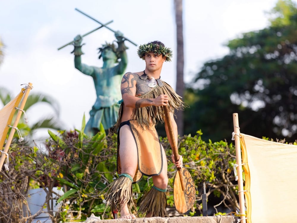 a man wearing traditional luau outfit holding a paddle by his side stands on stage in front of statue in background