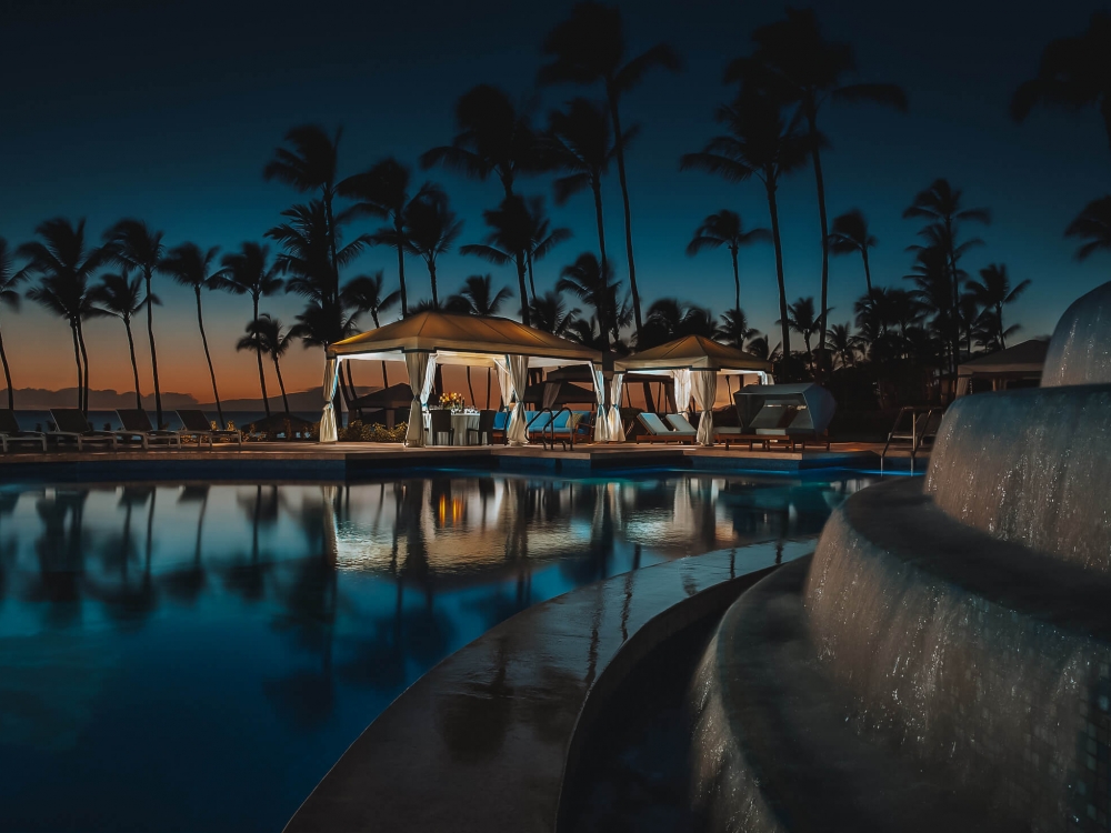 a water feature with multiple levels pours water into a larger pool with palm trees and cabanas in the background during sunset