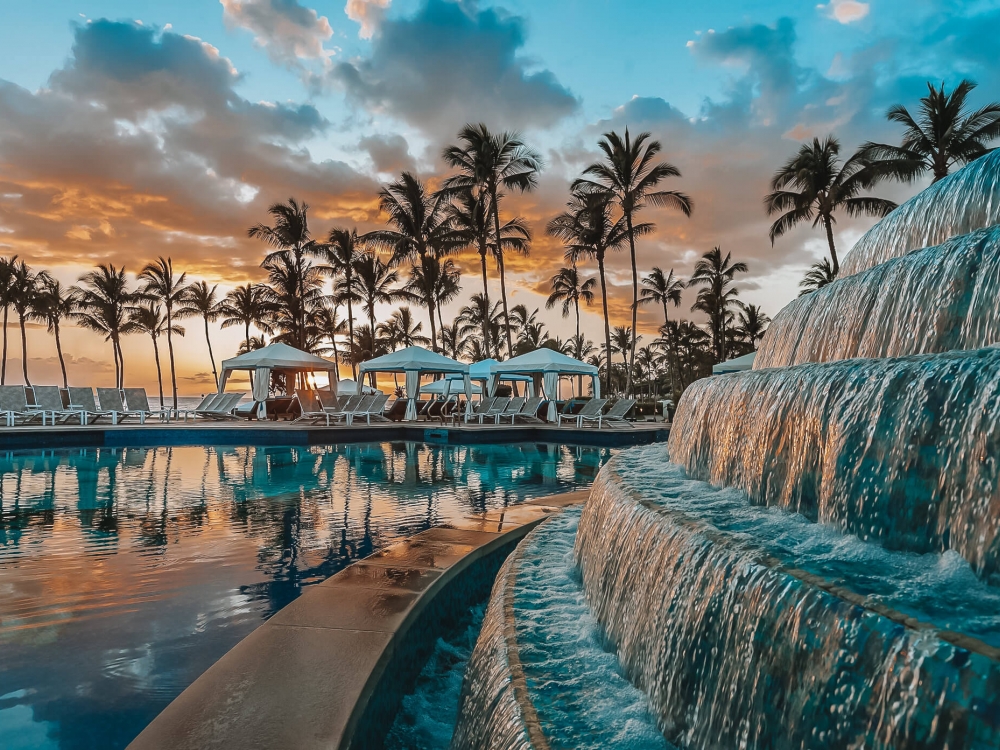 a water feature with multiple levels pours water into a larger pool with palm trees in the background at sunset