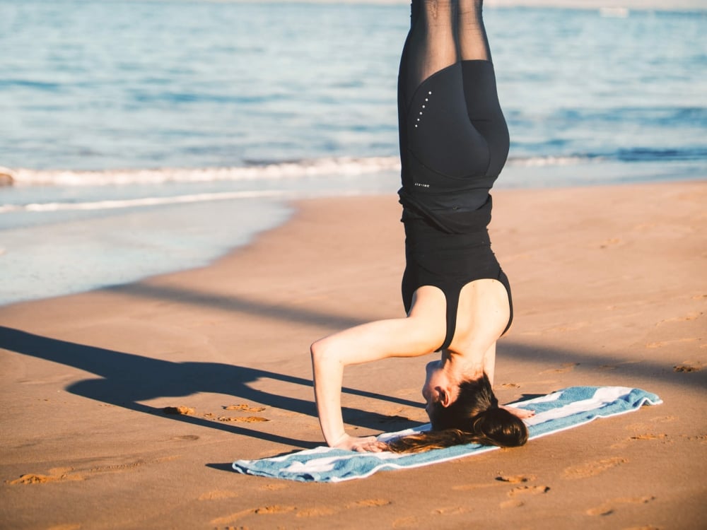 a woman in black workout clothing is in a headstand on a blue blanket on the beach beside the ocean