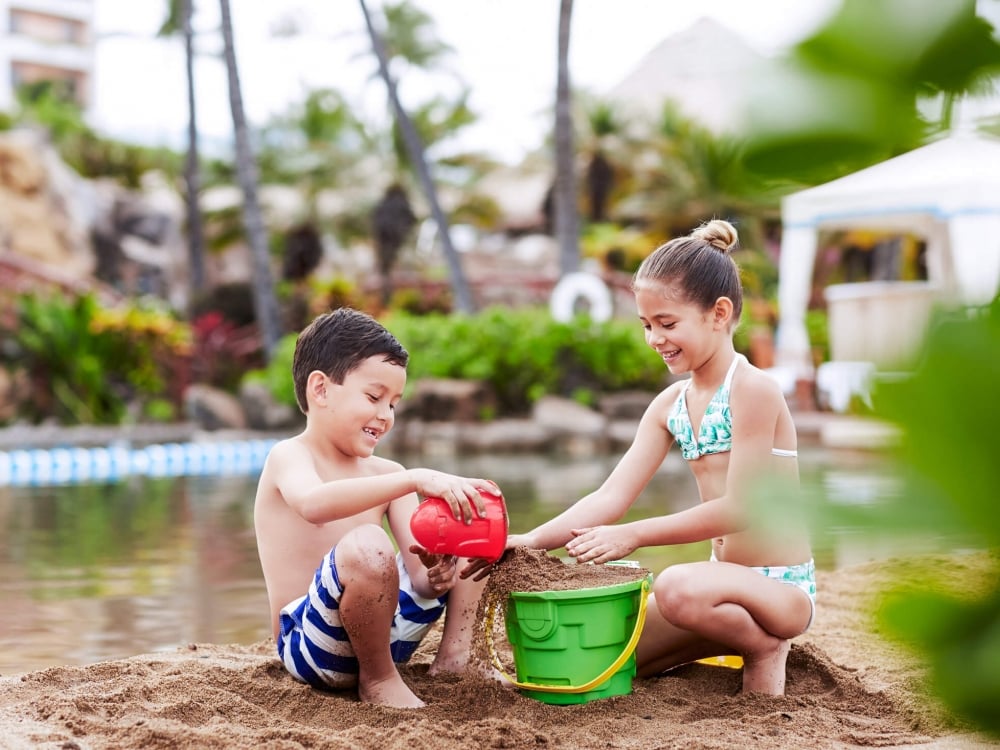 two kids play in the sand with green and red bucket with water in the background