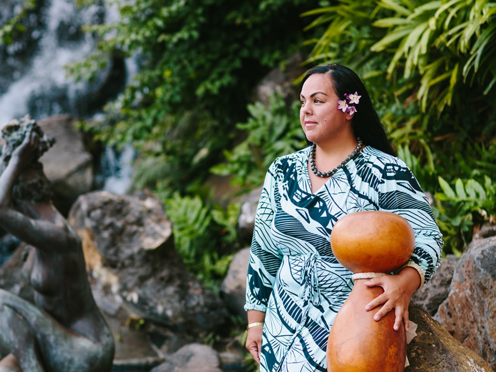 a woman in a blue striped dress leans against a rock near a waterfall holding a gourd