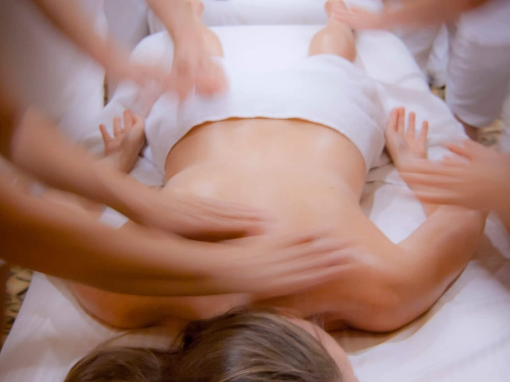 two people lay on their stomachs under white blankets while multiple massage therapists massage their bodies