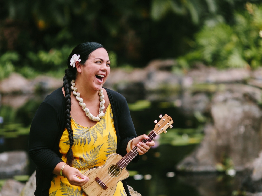 a woman in a yellow dress sits against a rock while strumming a ukulele and sings