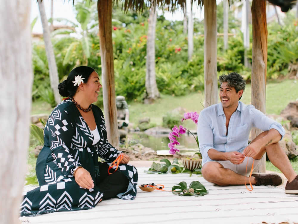 two people sit on white exterior platform surrounded by leaves and weavings