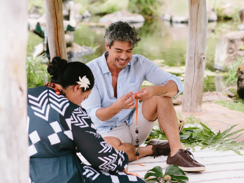 two people sit with piles of leaves at their feet while they weave the leaves with their hands