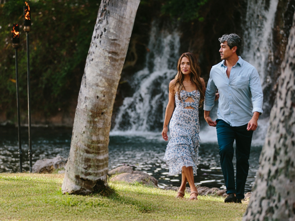 a man in a blue shirt and a woman in a strapless dress walk between two trees with a small waterfall feature and pond behind them