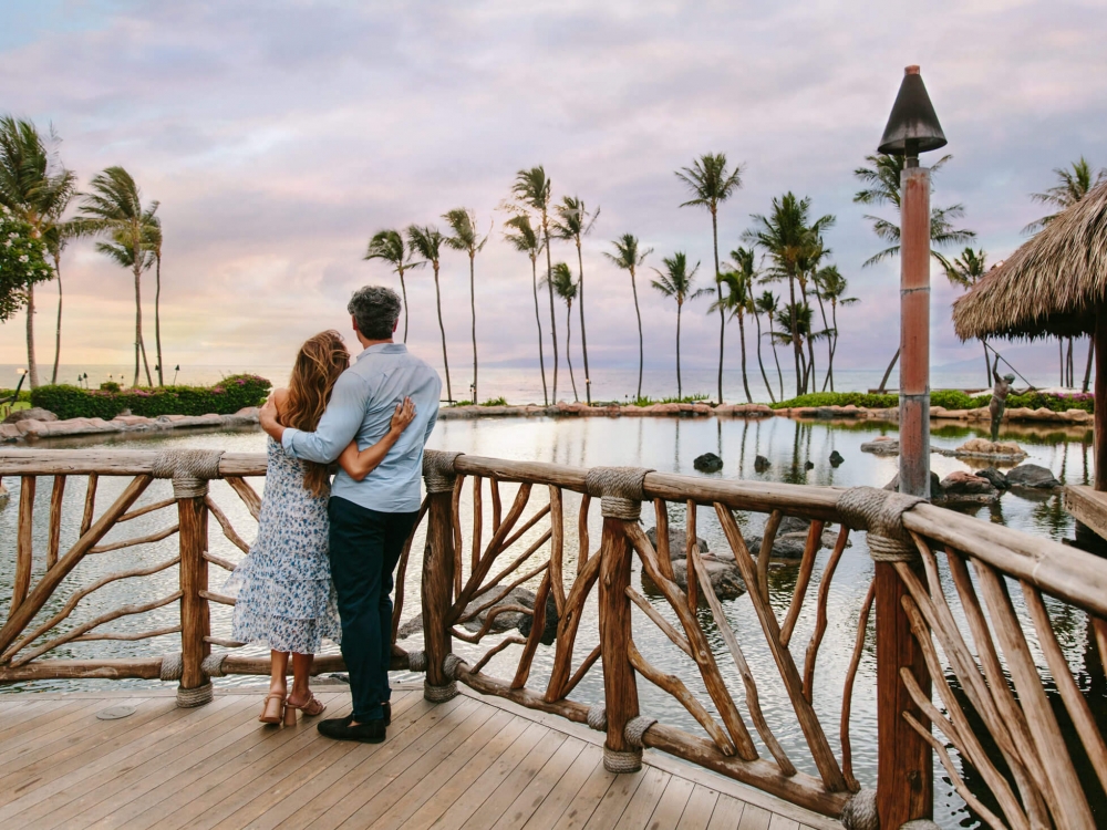 a couple stands on a wooden balcony while looking out at the sun setting