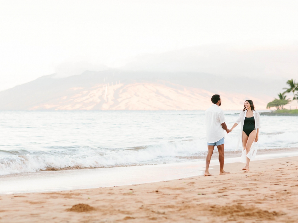 a couple walks along the beach beside the ocean with mountains in the background