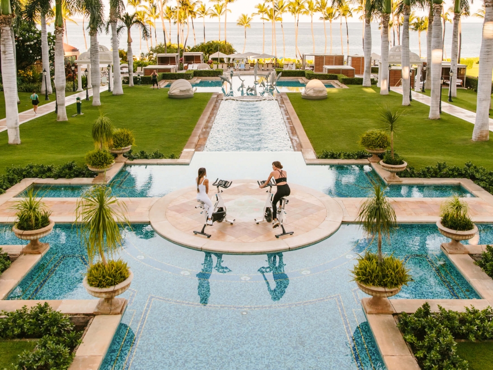 two women wearing black and white workout clothing ride on exercise bikes on platform between pools 