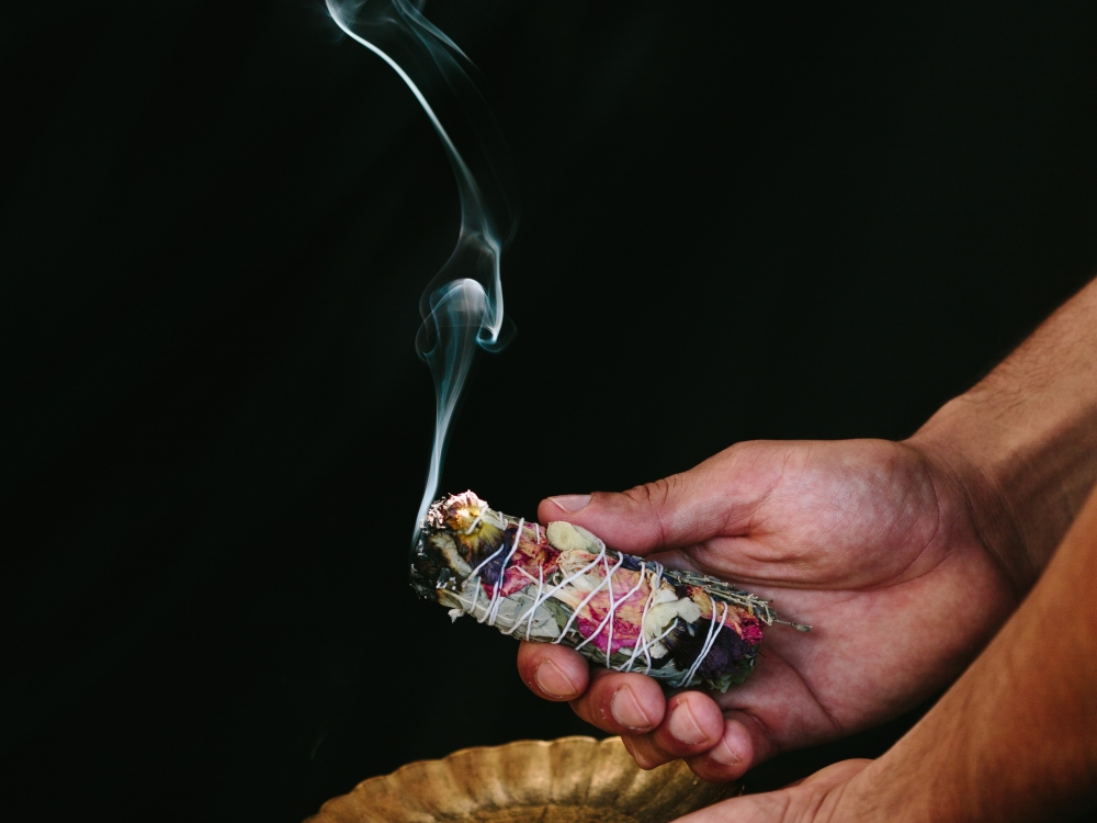 outheld hands hold a smoking bundle of herbs against a black backdrop