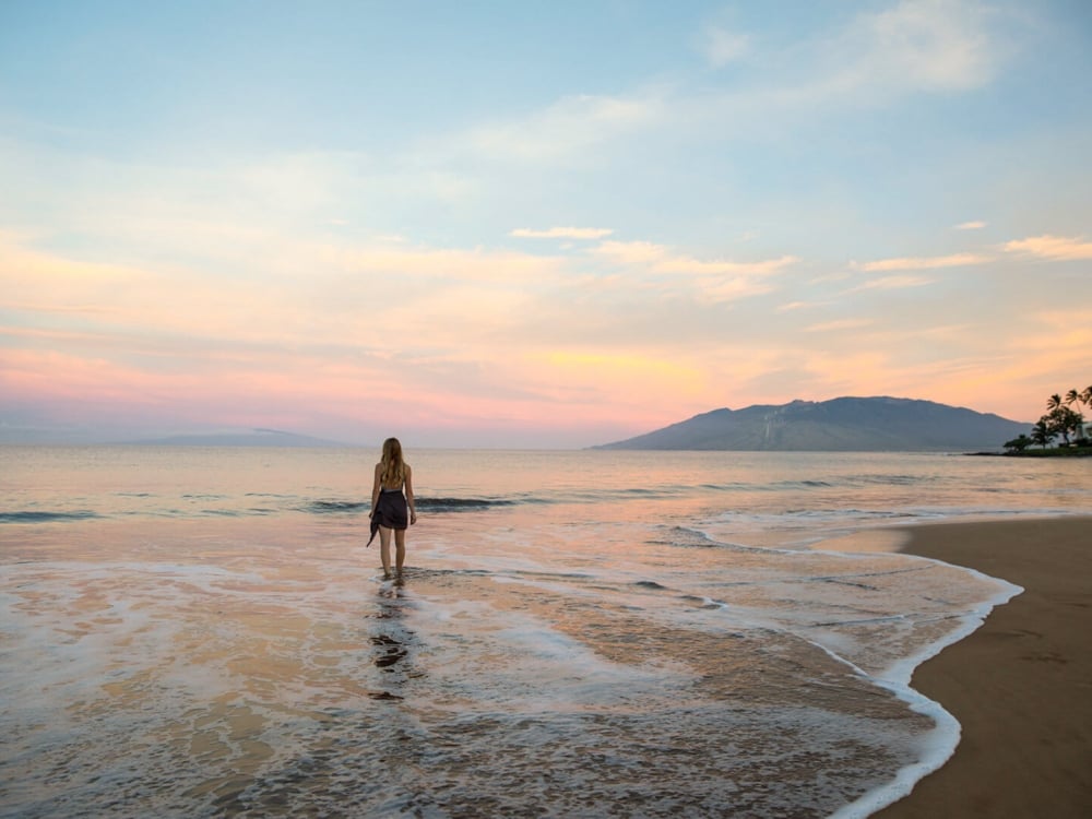a woman walks toward the ocean on the beach with mountains in the background while the sun sets