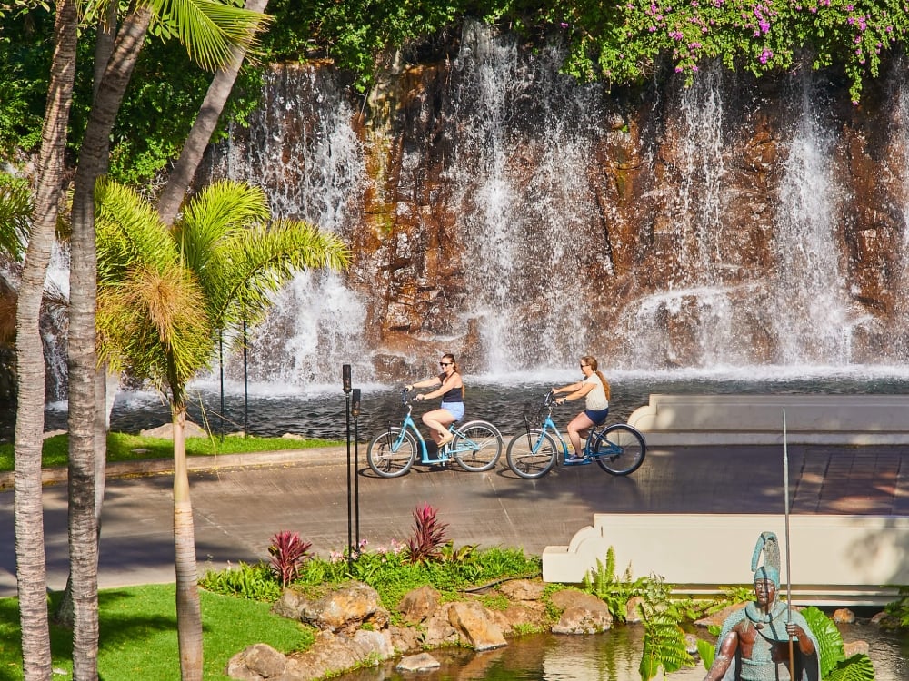 two people ride beach cruiser bikes along a path beside a wall of water