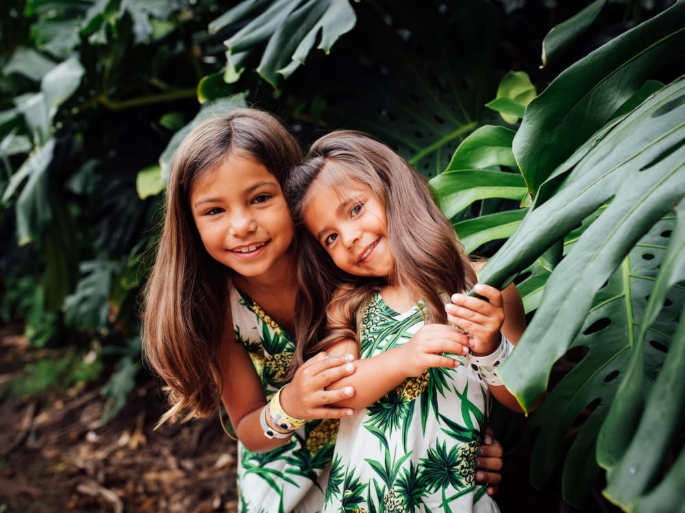 two small children wearing matching green and white outfits stand amongst green leaves of trees 