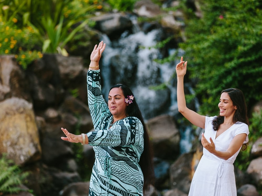 two women hold one hand above their head and one in front of them as they practice dancing in front of.a waterfall feature surrounded by foliage
