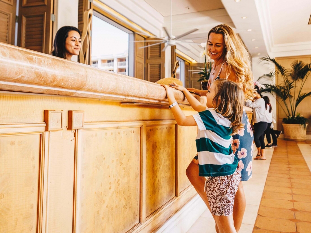 a woman and a child stand on one side of a wooden front desk while the child stands on their toes to see the person on the other side of the desk