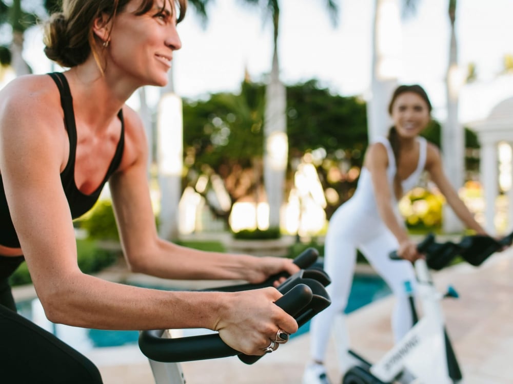 two women wearing black and white workout clothing ride on exercise bikes outside