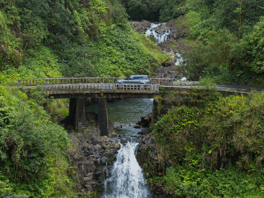 a vehicle drives across a bridge that's situated above a small waterfall and surrounded by green foliage and trees