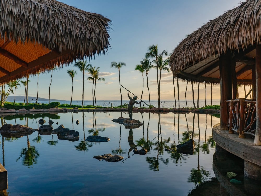 a statue holding a spear stands in the middle of a pond while the sun sets with a hut on either side with blue sky and palm trees in the background