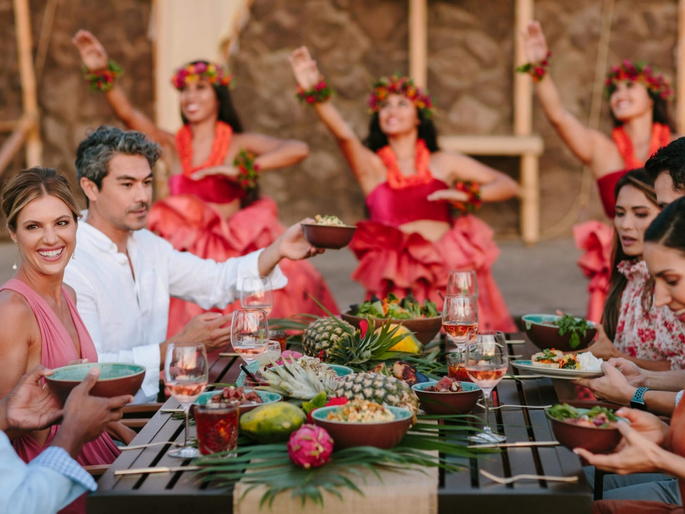 group of people enjoy dinner while women in red perform luau in the background