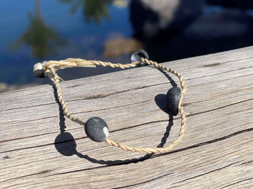 a small woven bracielet with two silver pieces sits on the edge of a wooden dock with palm trees reflected off the water in the background