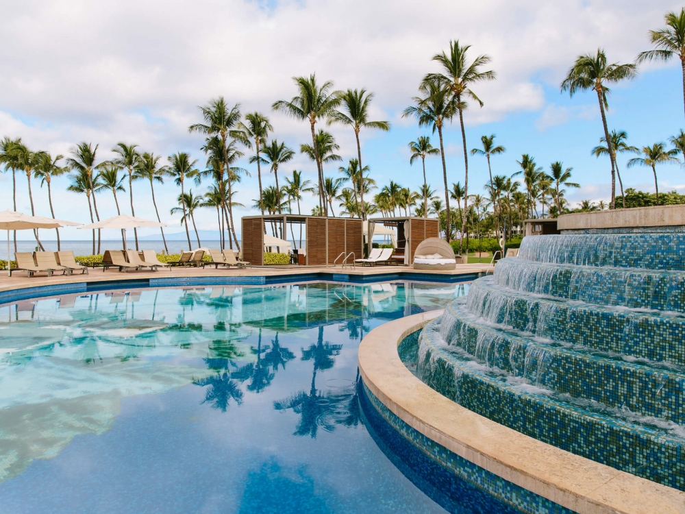 a water feature with multiple levels pours water into a larger pool with palm trees in the background