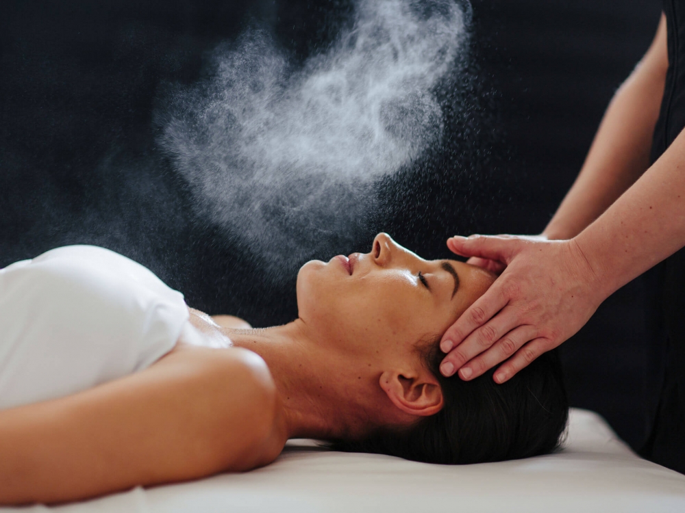 woman lays on her back under a white blanked while her head is massaged and mist is sprayed over her face
