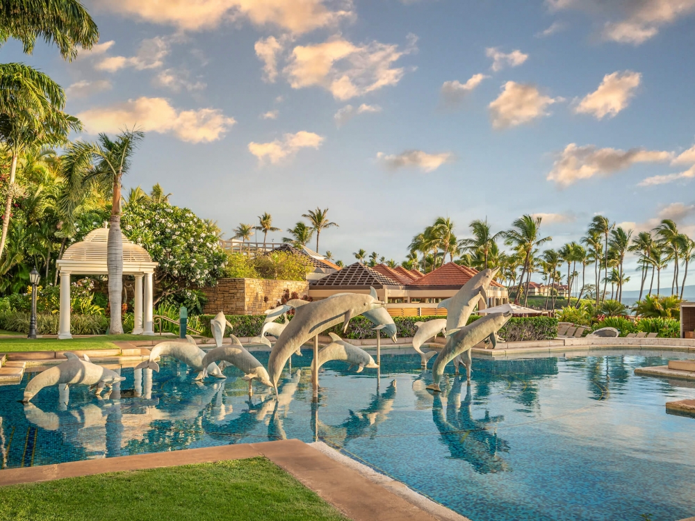 group of stone dolphin statues jump out of a blue pool surrounded by trees and with clouds in the sky