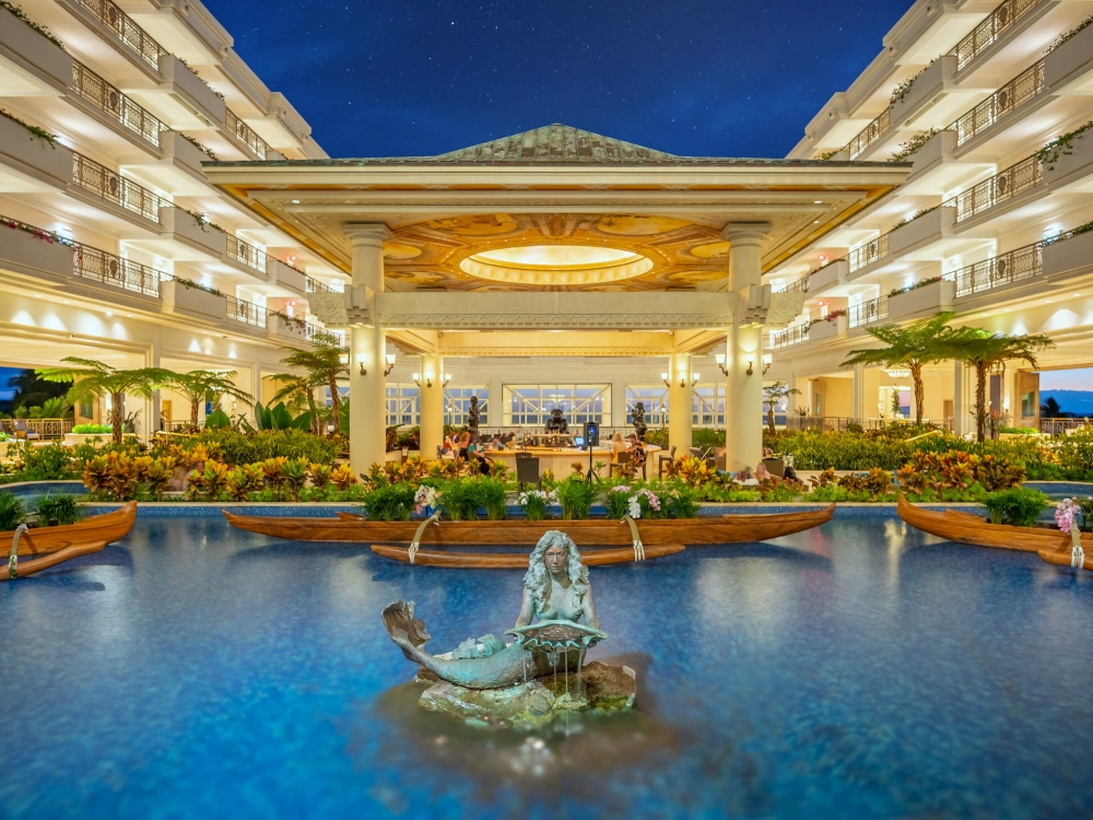 a stone mermaid statue sits in the center of a blue pool with lit up building in the background