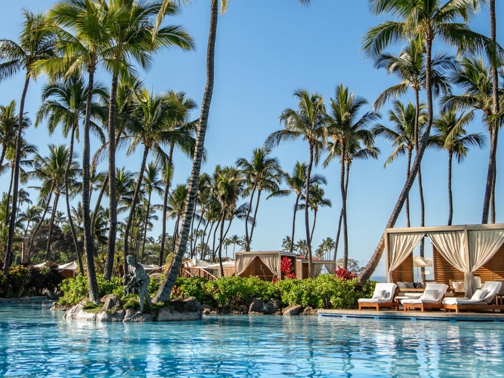 Sun falls on a sparkling blue pool lined with luxury cabanas and waving palm trees