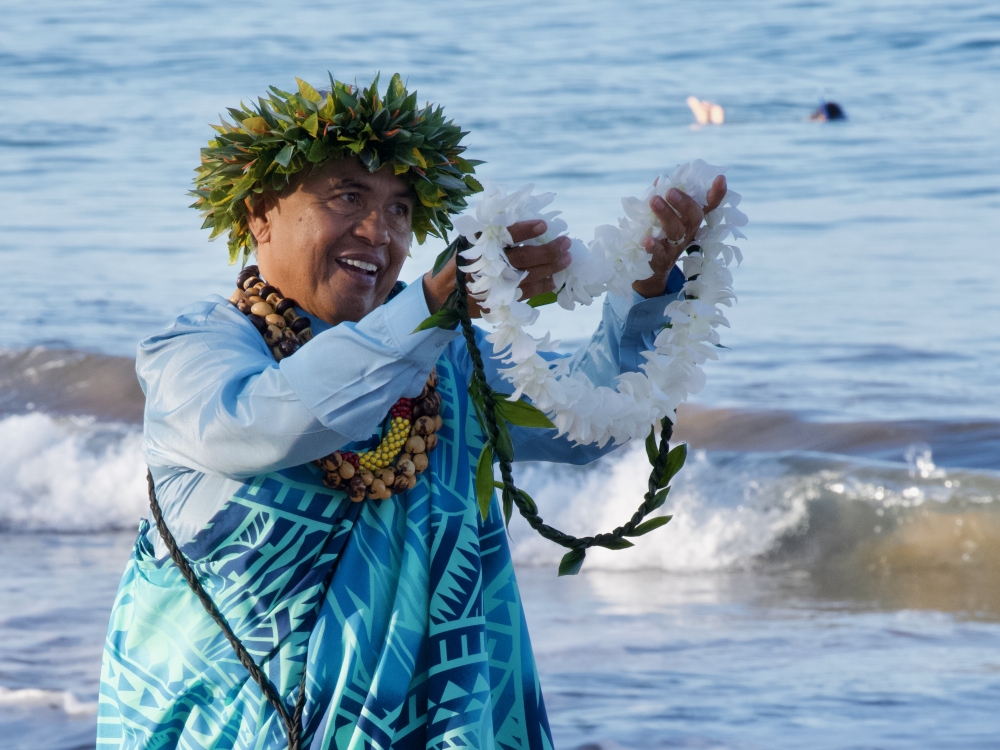 A Hawaiian man on a beach holding out a lei