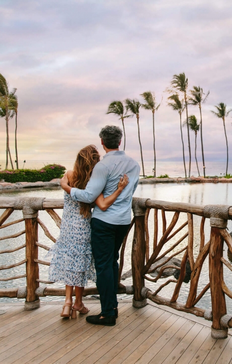 a couple stands on a wooden balcony while looking out at the sun setting