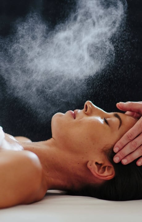 woman lays on her back under a white blanked while her head is massaged and mist is sprayed over her face