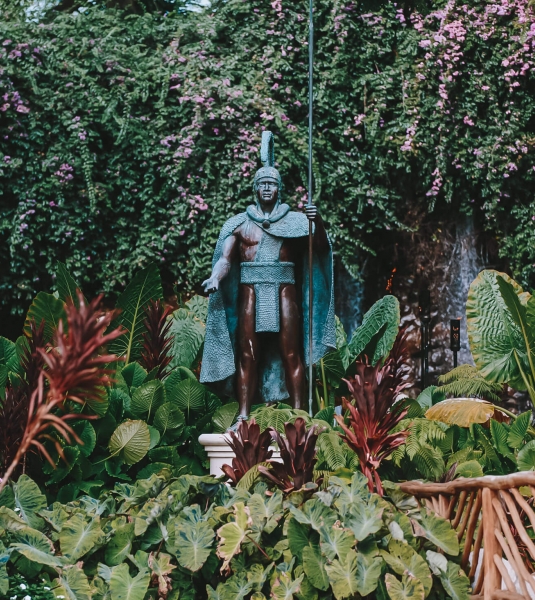 a statue of warrior stands amongst green foliage in front of a forest of trees