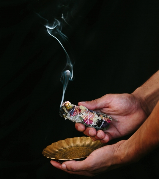 outheld hands hold a smoking bundle of herbs and a small copper tray against a black backdrop