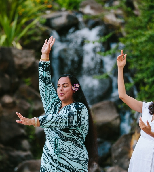 two women hold one hand above their head and one in front of them as they practice dancing in front of.a waterfall feature surrounded by foliage