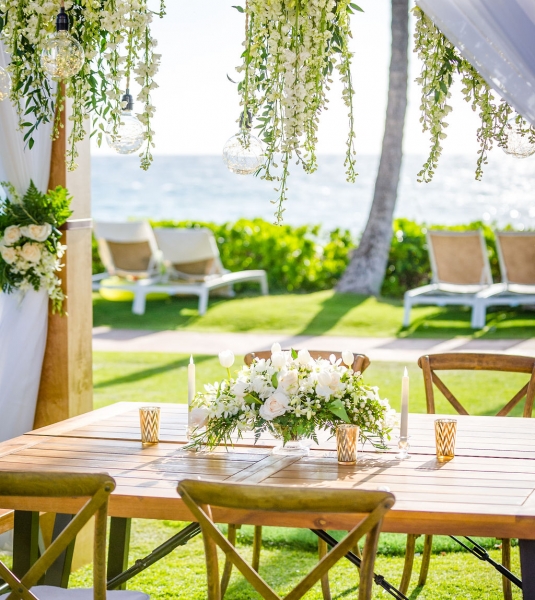 chairs sit around wooden table with floral centerpiece under an awning with trailing greenery and flowers hang from the edge