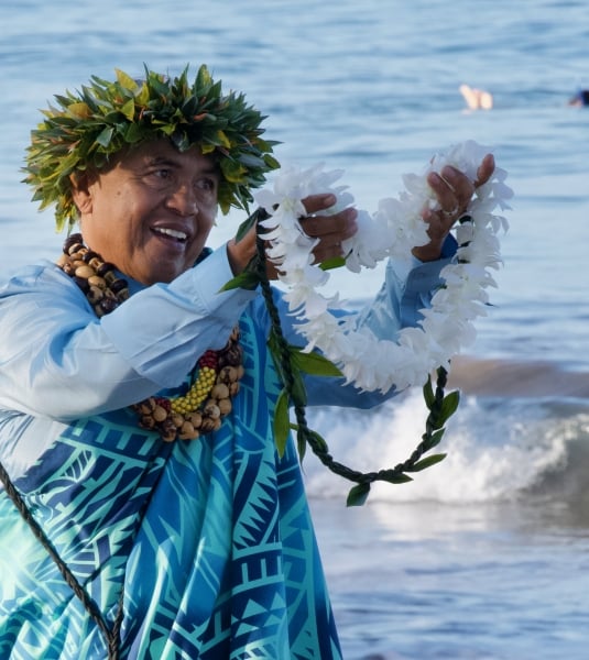 A Hawaiian man on a beach holding out a lei
