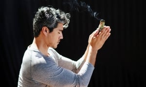 side profile of a man sitting crosslegged on the ground holds his hands up while smoke trails over his head