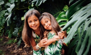 two small children wearing matching green and white outfits stand amongst green leaves of trees 