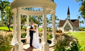 bride and groom stand under a gazebo with a chapel in the background