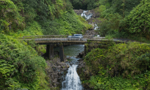 a vehicle drives across a bridge that's situated above a small waterfall and surrounded by green foliage and trees