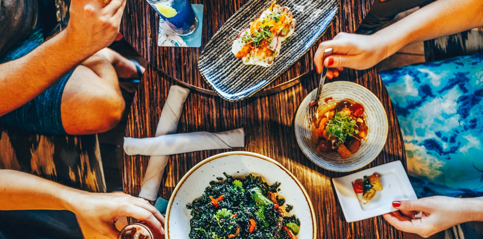 multiple plates of food sit on a wooden table while four people reach over the food with their utensils