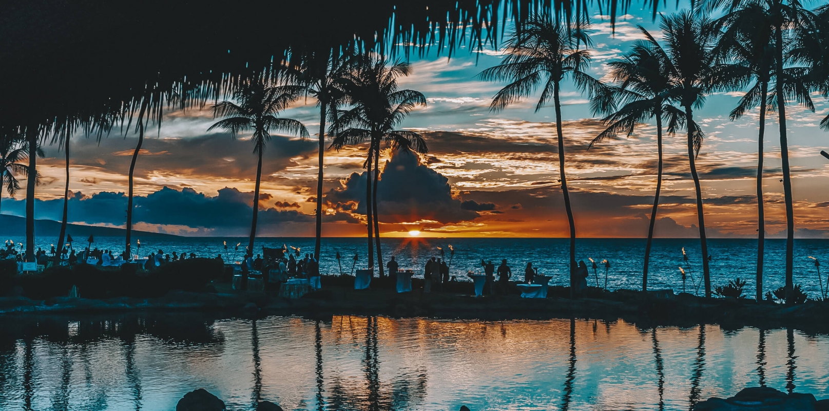 shadow of palm trees reflect off the water in the foreground while the sun sets over the ocean creating an orange and blue sky